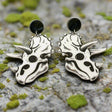 A pair of triceratops skull acrylic earrings in black and white with round black acrylic toppers against a grey background.