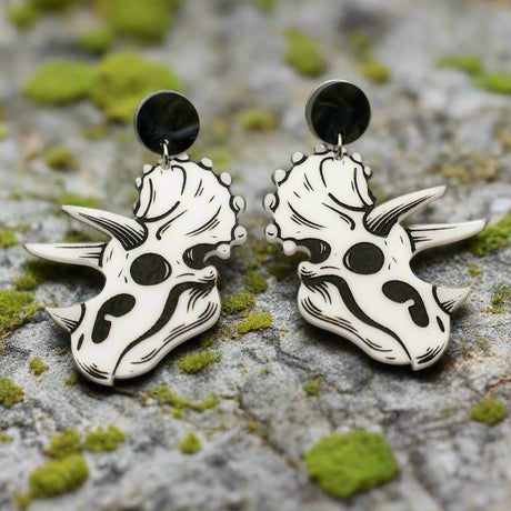 A pair of triceratops skull acrylic earrings in black and white with round black acrylic toppers against a grey background.