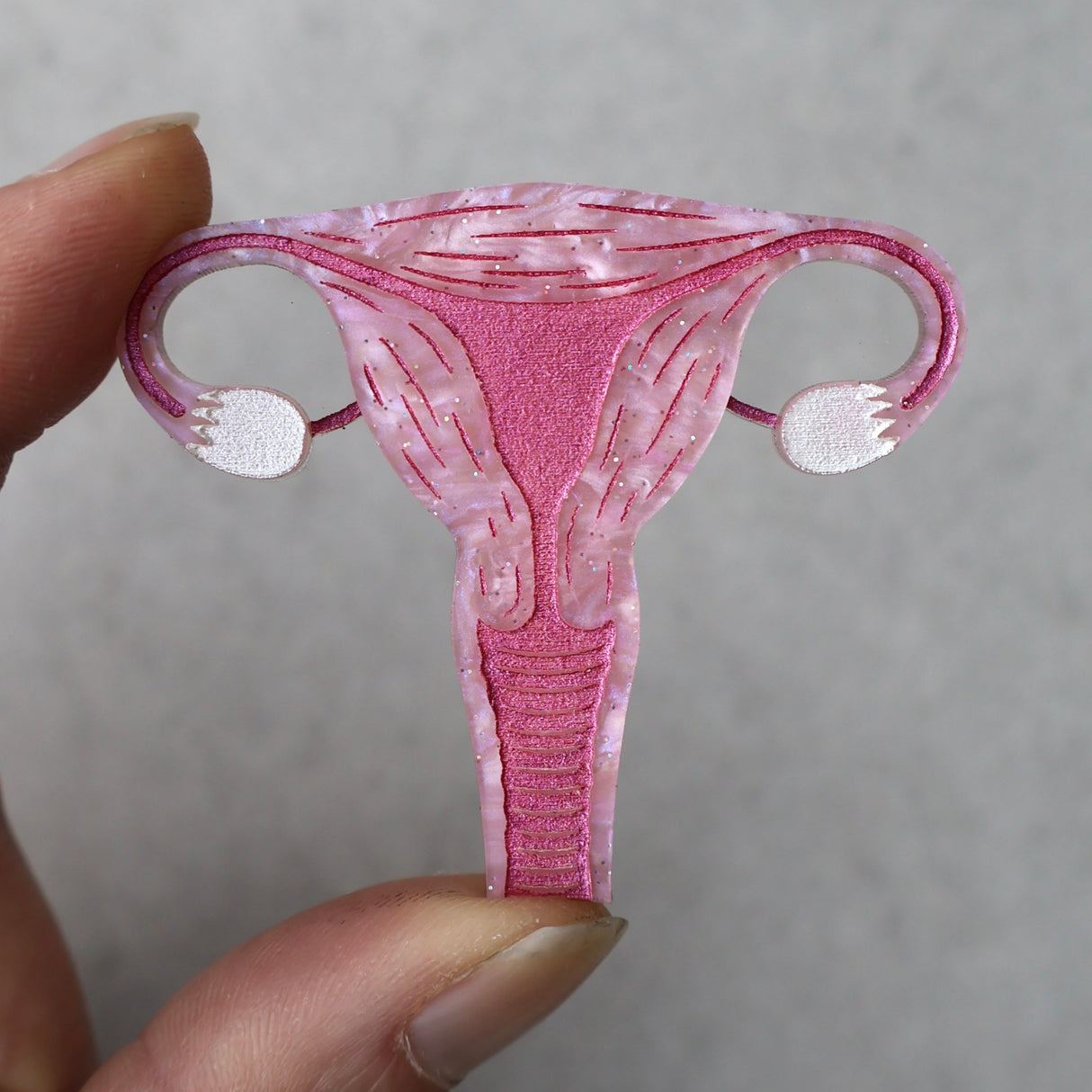 A closeup view of a laser cut acrylic uterus brooch, in pink acrylic with pink painted details, being held up by a hand against a grey background.