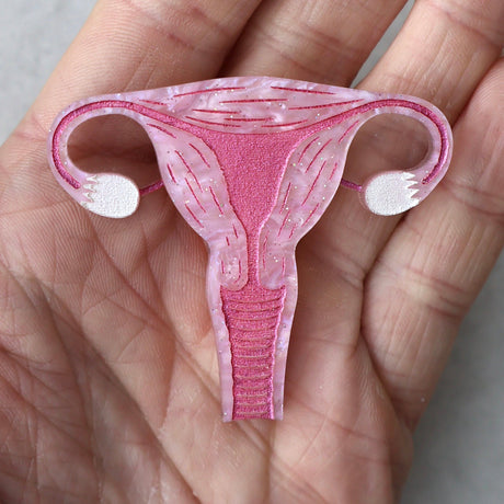 A closeup view of a laser cut acrylic uterus brooch, in pink acrylic with pink painted details being held in a pam.