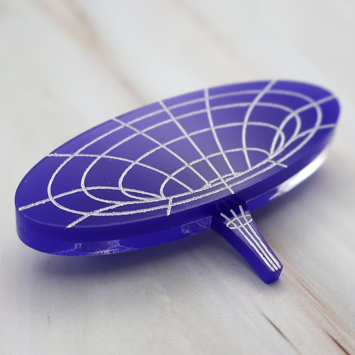 Angled view of dark blue wormhole brooch made from laser cut acrylic with hand painted details, depicting the warping of spacetime