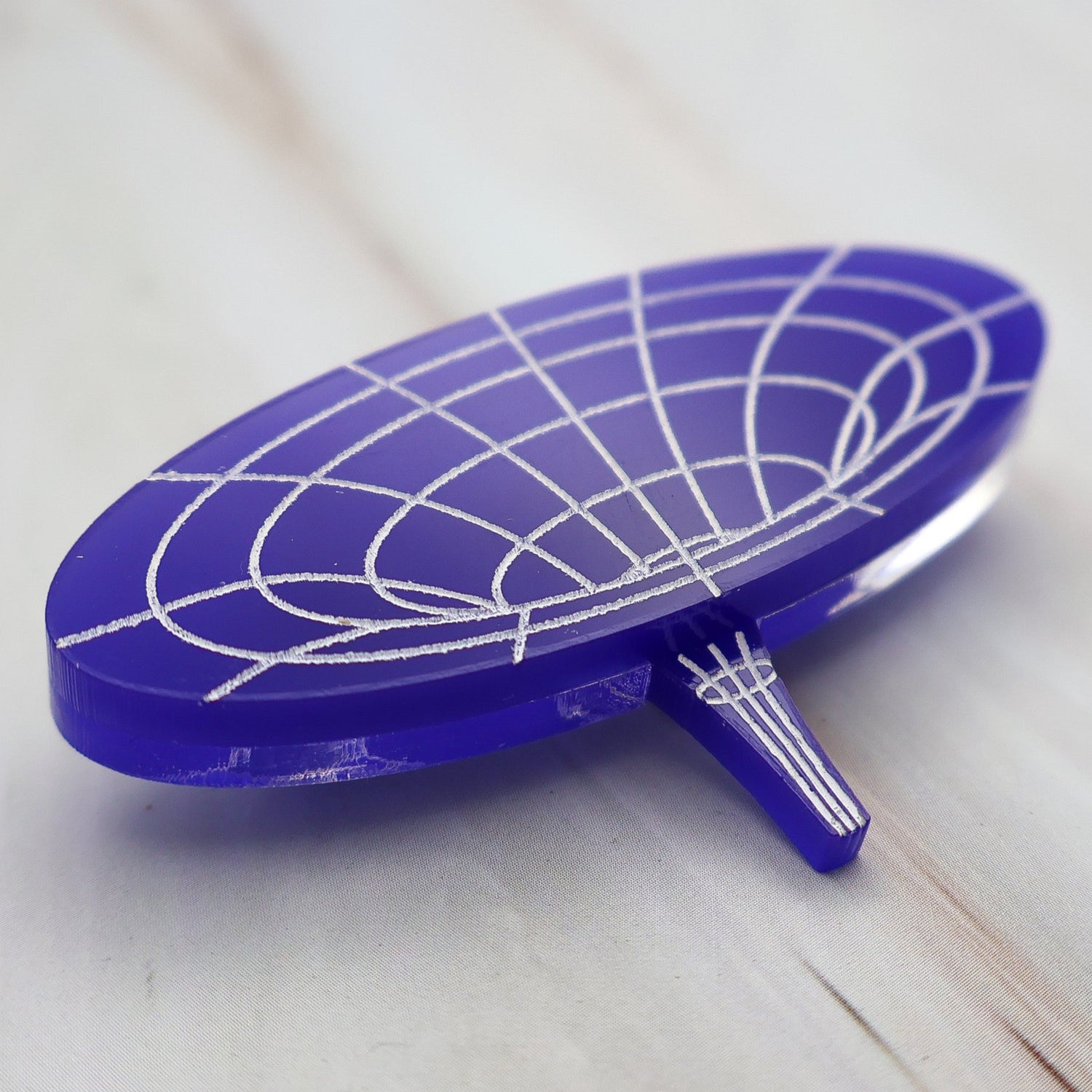 Angled view of dark blue wormhole brooch made from laser cut acrylic with hand painted details, depicting the warping of spacetime