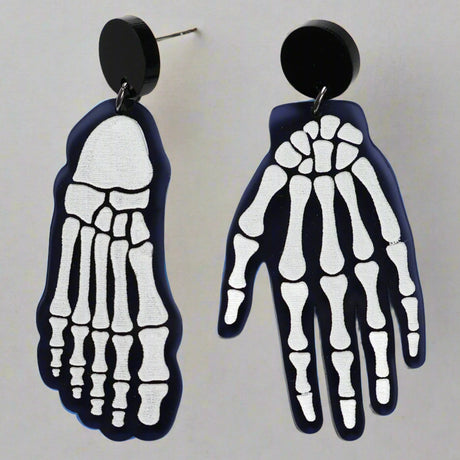 A pair of mismatched earrings, one in the shape of a hand and the other a foot, with a x-ray bone pattern on a black background.