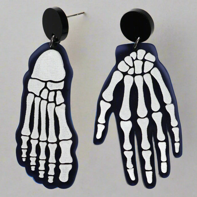 A pair of mismatched earrings, one in the shape of a hand and the other a foot, with a x-ray bone pattern on a black background.