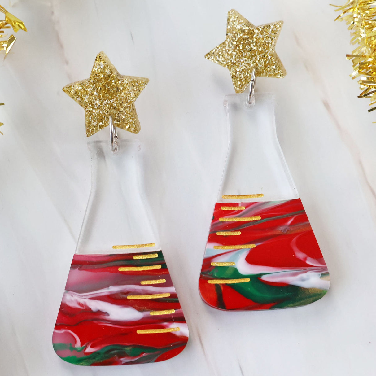 A pair of dangle earrings featuring a conical flask design with Christmas-themed decorations, including red and green swirls and gold accents, topped with glittery star-shaped acrylics.