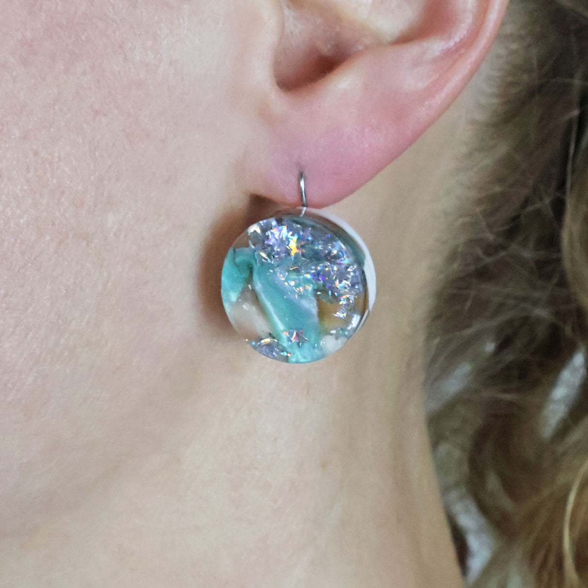 A pair of one of a kind round recycled acrylic earrings with stainless steel french hooks and green silver and brown acrylics being modelled on an ear.