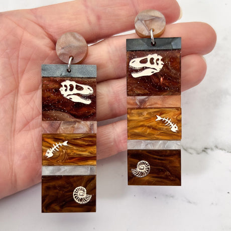 Dangle earrings representing sedimentary layers, comprising 6 strips of different grey and brown acrylics, with a t-rex skull, fish bones and nautilus engraved and handpainted in the larger layers. They hang from brown toppers and are being held up.