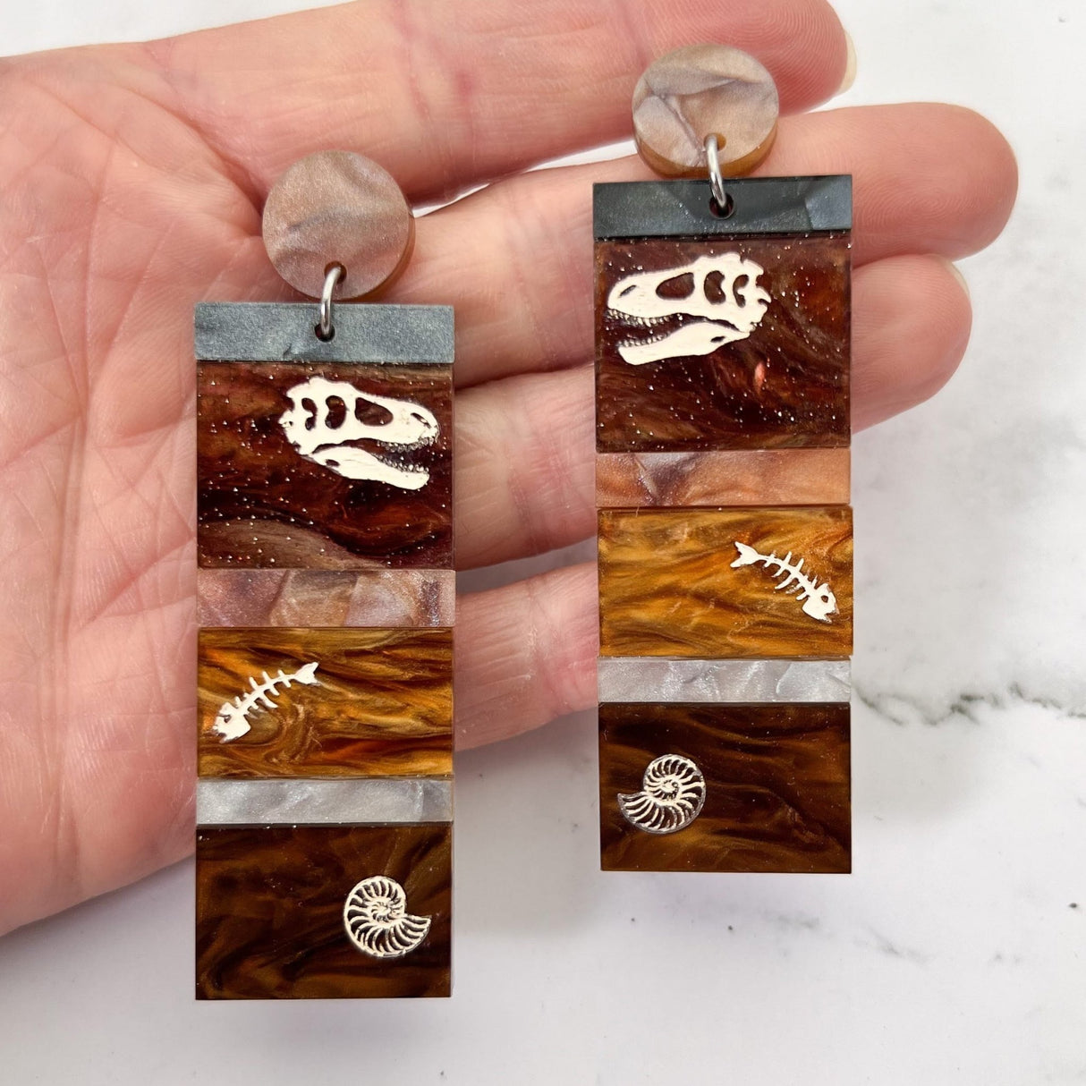 Dangle earrings representing sedimentary layers, comprising 6 strips of different grey and brown acrylics, with a t-rex skull, fish bones and nautilus engraved and handpainted in the larger layers. They hang from brown toppers and are being held up.