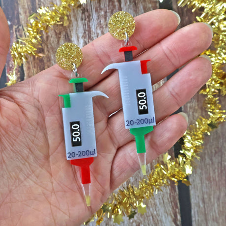 Earrings shaped like laboratory micropipettes in Christmas colours, held in a hand with tinsel in the background.