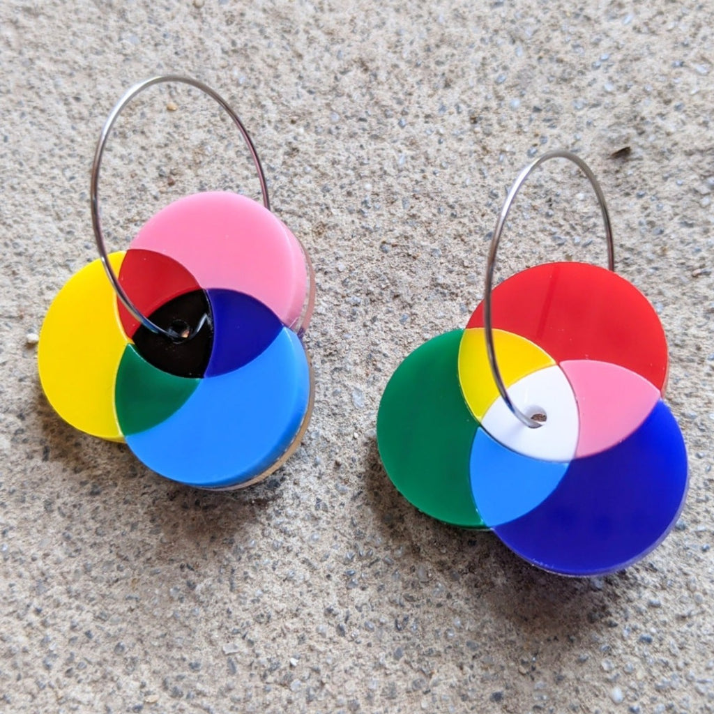 Asymmetrical Laser Cut Acrylic Colour Wheel Earrings showing additive and subtractive colour diagrams. Earrings are sitting on a concrete surface.