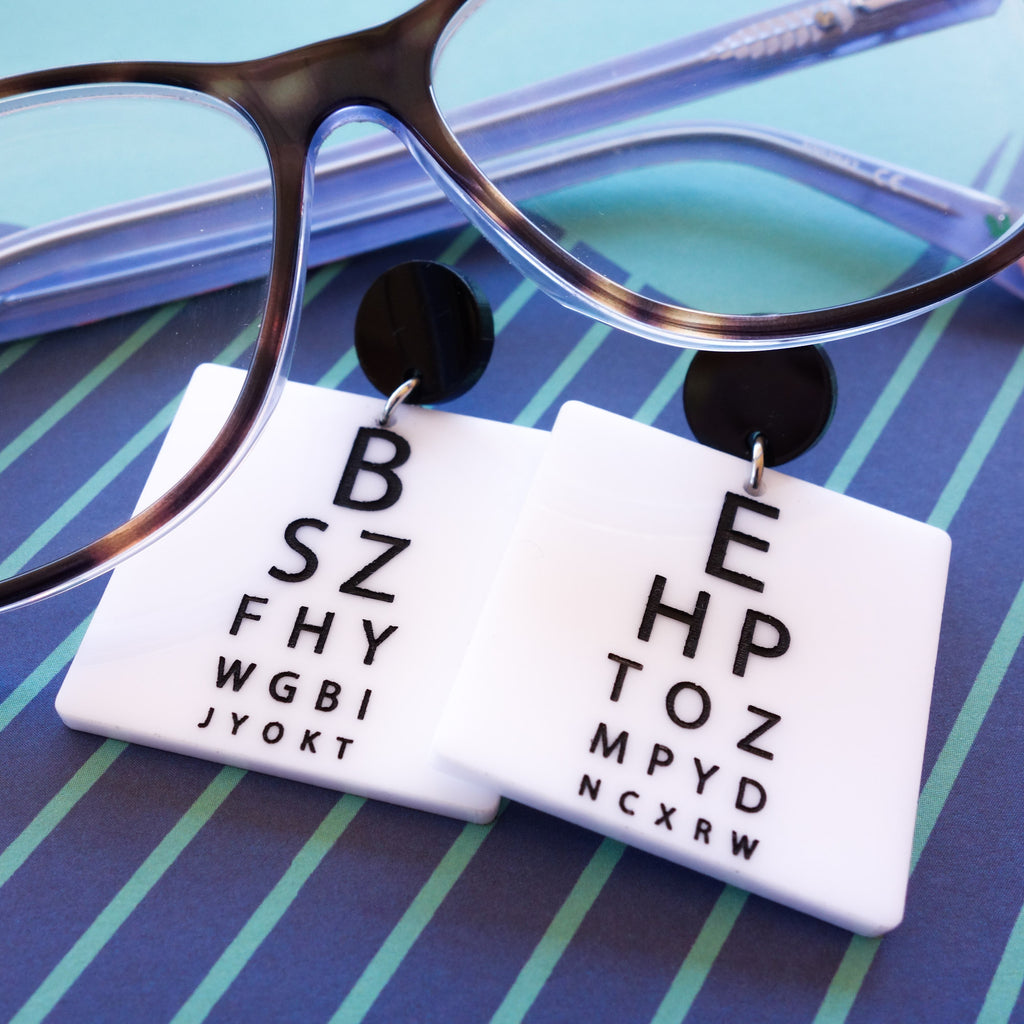 Laser cut acrylic eye chart earrings. With black text of a white background, and with black earring toppers. a pair of glasses are included as a prop.