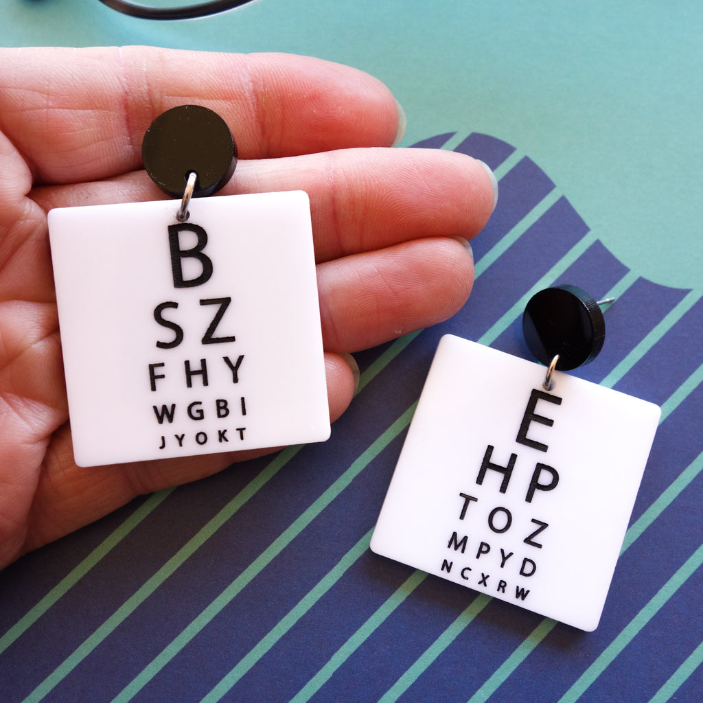 Laser cut acrylic eye chart earrings. With black text of a white background, and with black earring toppers. 