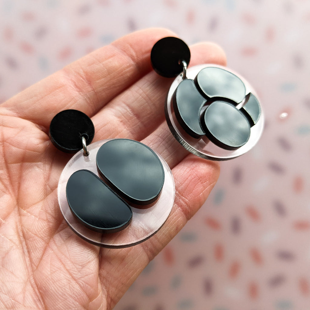 Asymmetrical meiosis earrings, with black shapes against clear circle backings, hanging from black earring toppers. Angled view in hands.