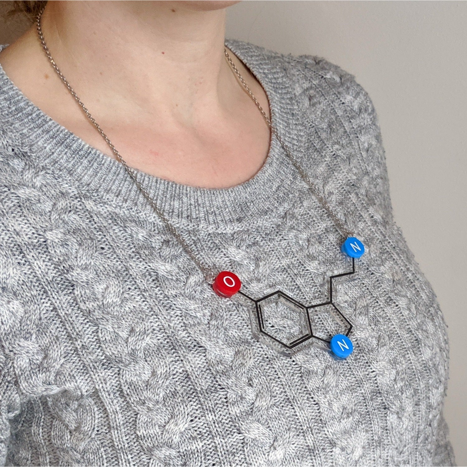 A person wearing a grey sweater is shown with a necklace featuring the serotonin molecule design. The necklace is composed of clear, red, and blue acrylic pieces representing the molecular structure, with black paint detailing the chemical bonds.