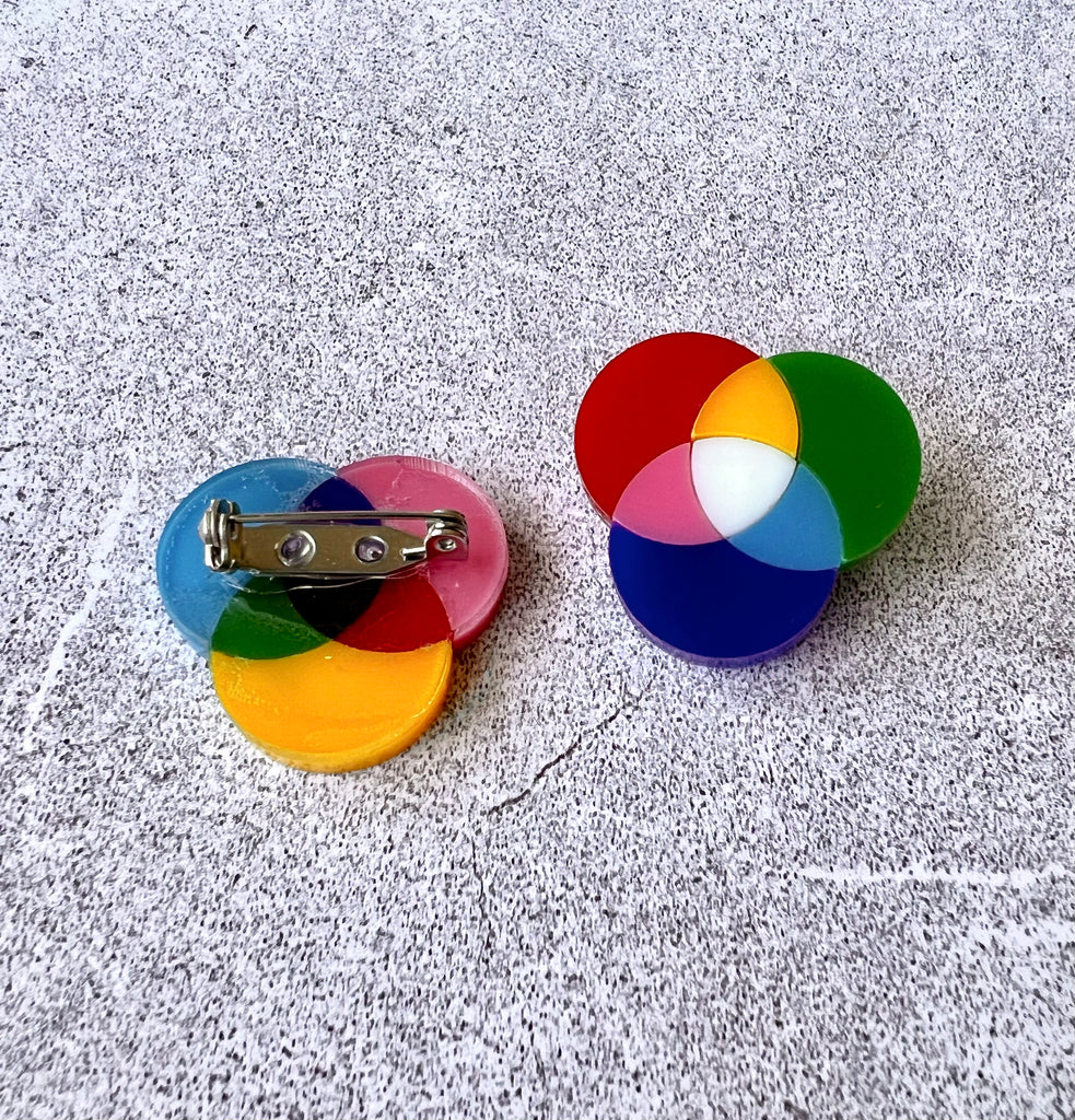 A pair of small subtractive and additive colour wheel brooches, laser cut in acrylic. The back of one brooch shows the stainless steel brooch pin with rolling clasp.