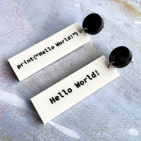 White dangle asymmetrical acrylic earrings with black text showing code to display Hello World. Hanging from black earring toppers .