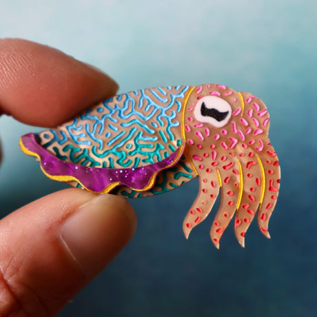 Colourful acrylic cuttlefish brooch held between fingers against a blue background