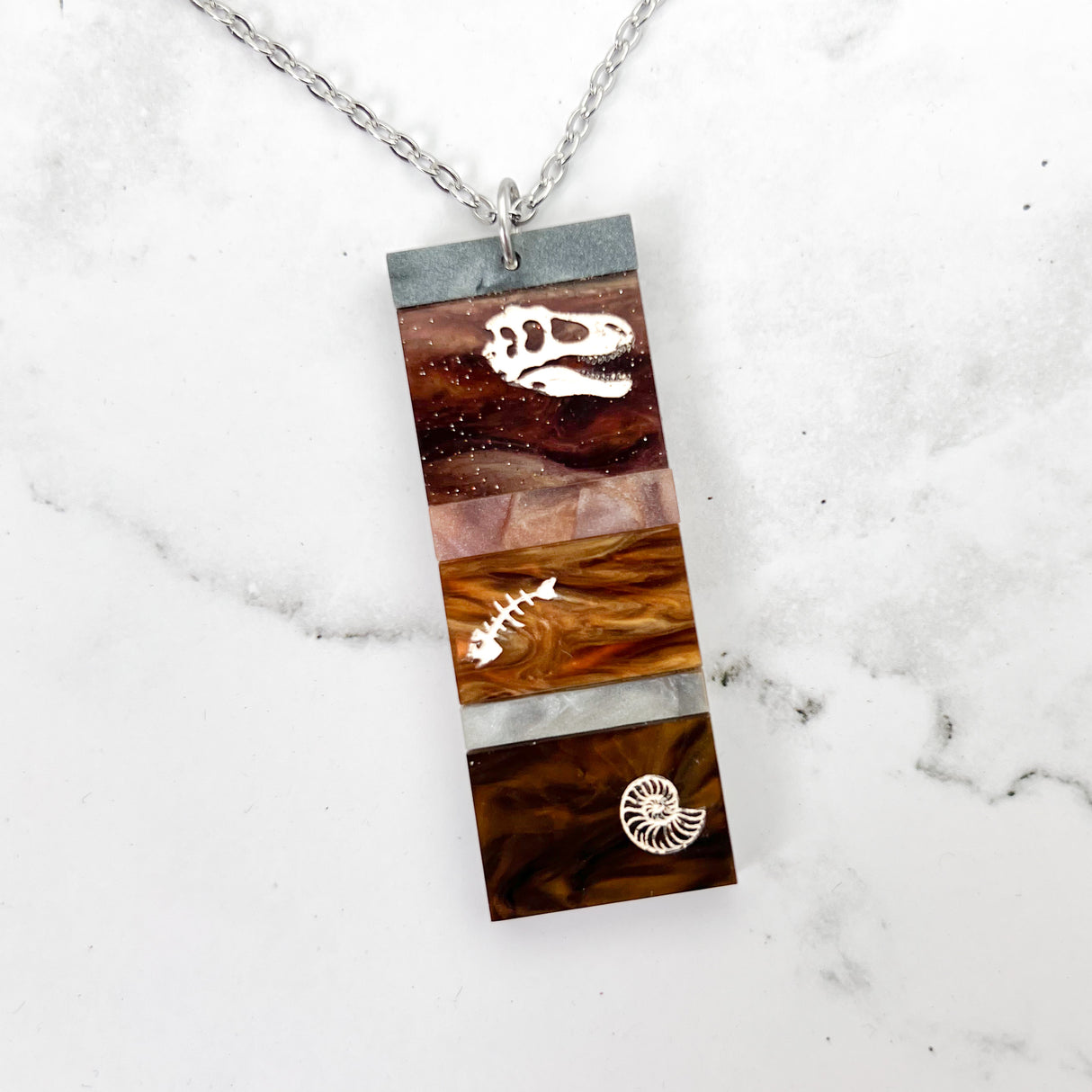 A pendant representing sedimentary layers, rectangular in shape and comprising 6 strips of different grey and brown acrylics, with a t-rex skull, fish bones and nautilus engraved and handpainted in the larger layers. The pendant hangs on a fine stainless steel chain.