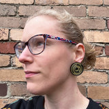 A pair of round star trail studs,black with gold painted star trails within. Studs are being modelled by the maker, a woman with blonde hair and glasses.