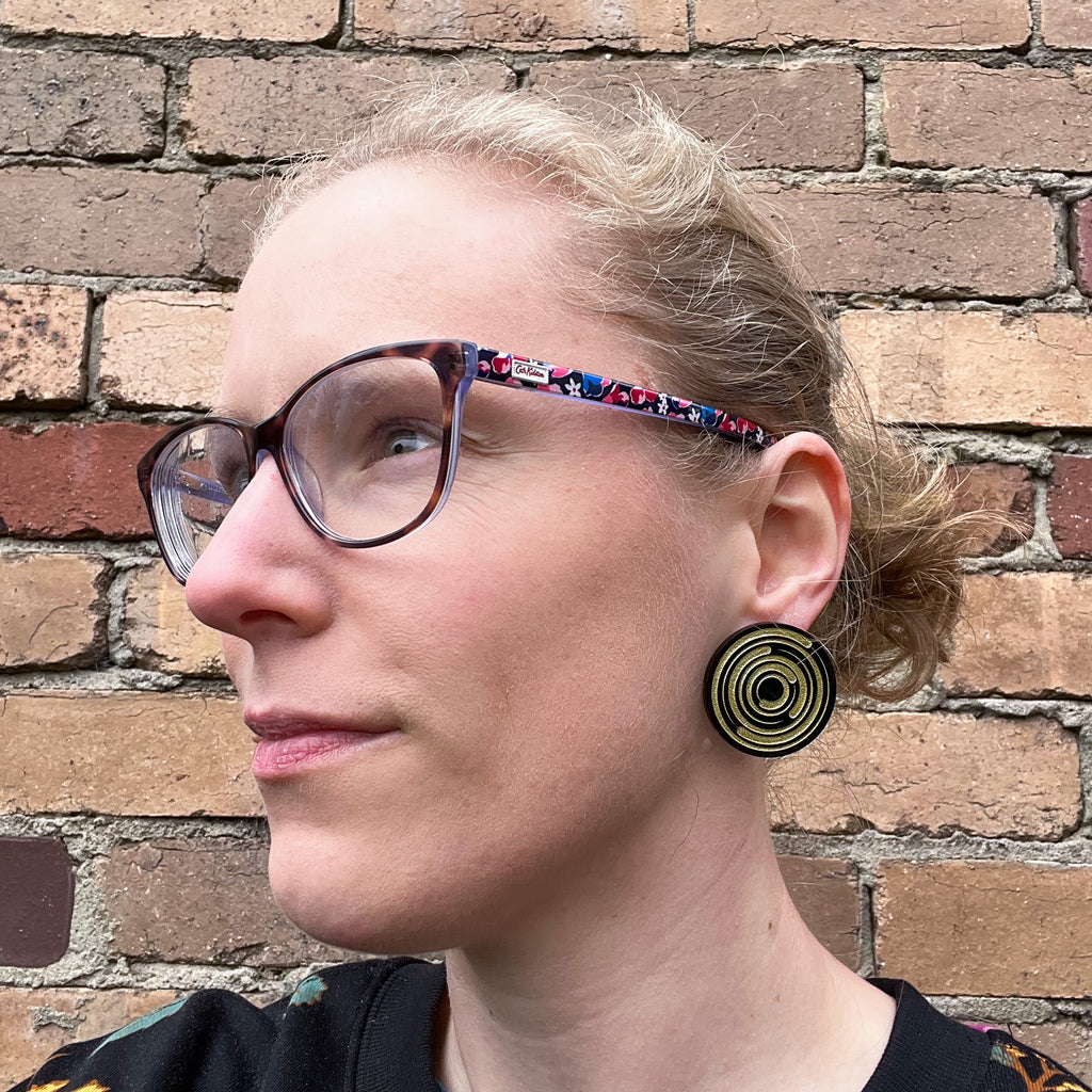A pair of round star trail studs,black with gold painted star trails within. Studs are being modelled by the maker, a woman with blonde hair and glasses.