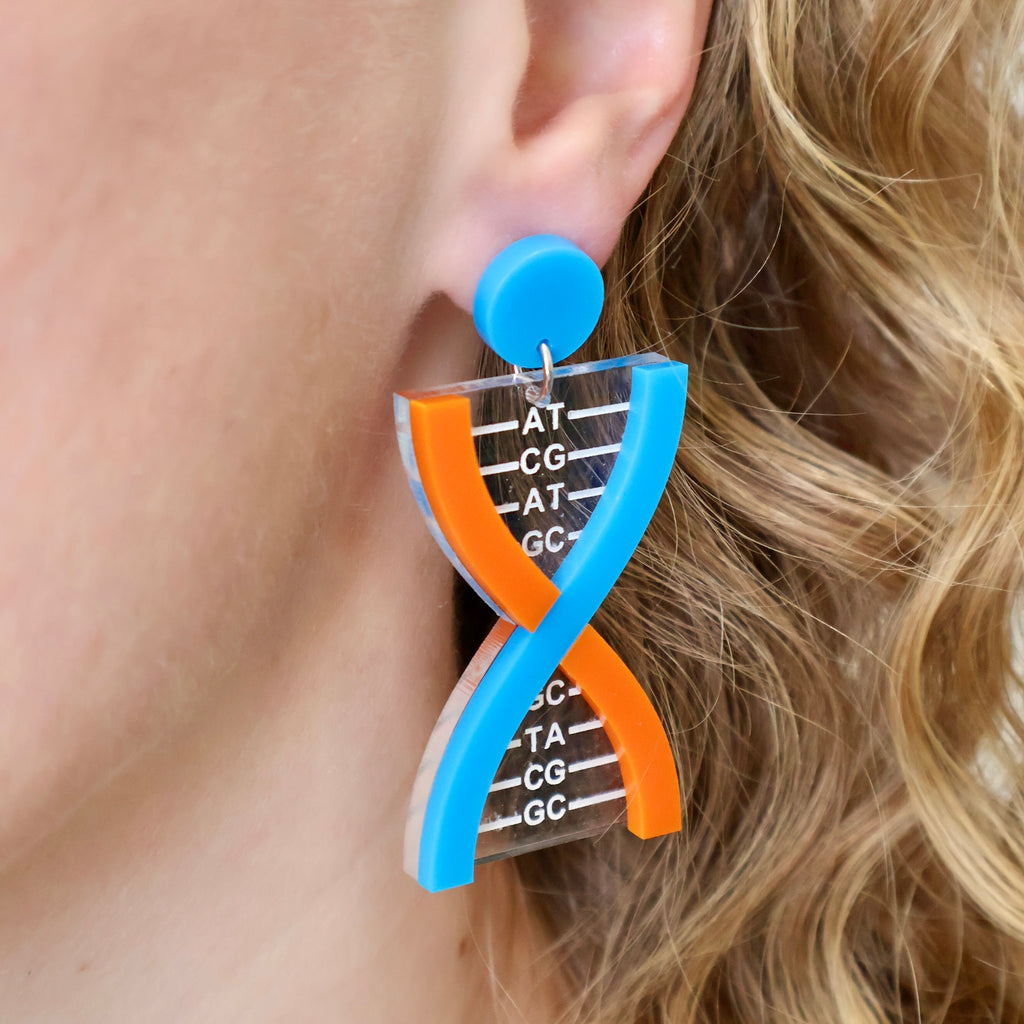 Laser cut acrylic DNA earrings being modelled. The colour scheme of the RNA backbones are orange and blue, and the base pairs are engraved and handpainted white on a clear acrylic. 