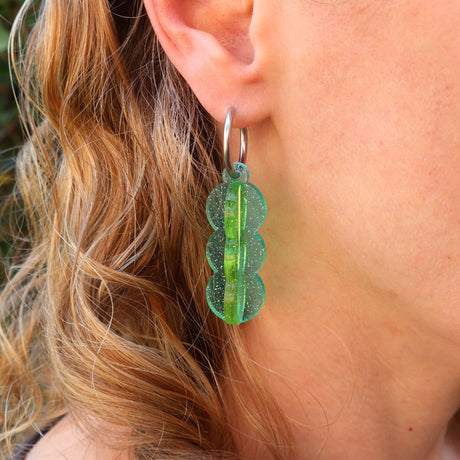 A pair of blue and green transparent acrylic earrings with a 3D construction and huggie hoops, being modelled in an ear. 
