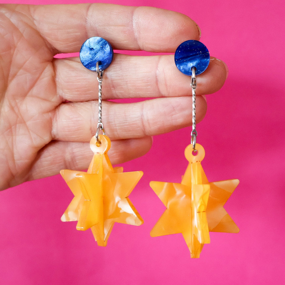 Hand holding a pair of earrings with blue tops and yellow star-shaped drops against a pink background