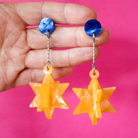 Hand holding a pair of earrings with blue tops and yellow star-shaped drops against a pink background