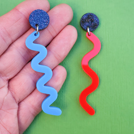 A pair of asymmetrical acrylic earrings with blue and red wavelength designs being held up by a hand on a green background