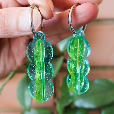 A pair of blue and green transparent acrylic earrings with a 3D construction and huggie hoops, being held up against a brick background.