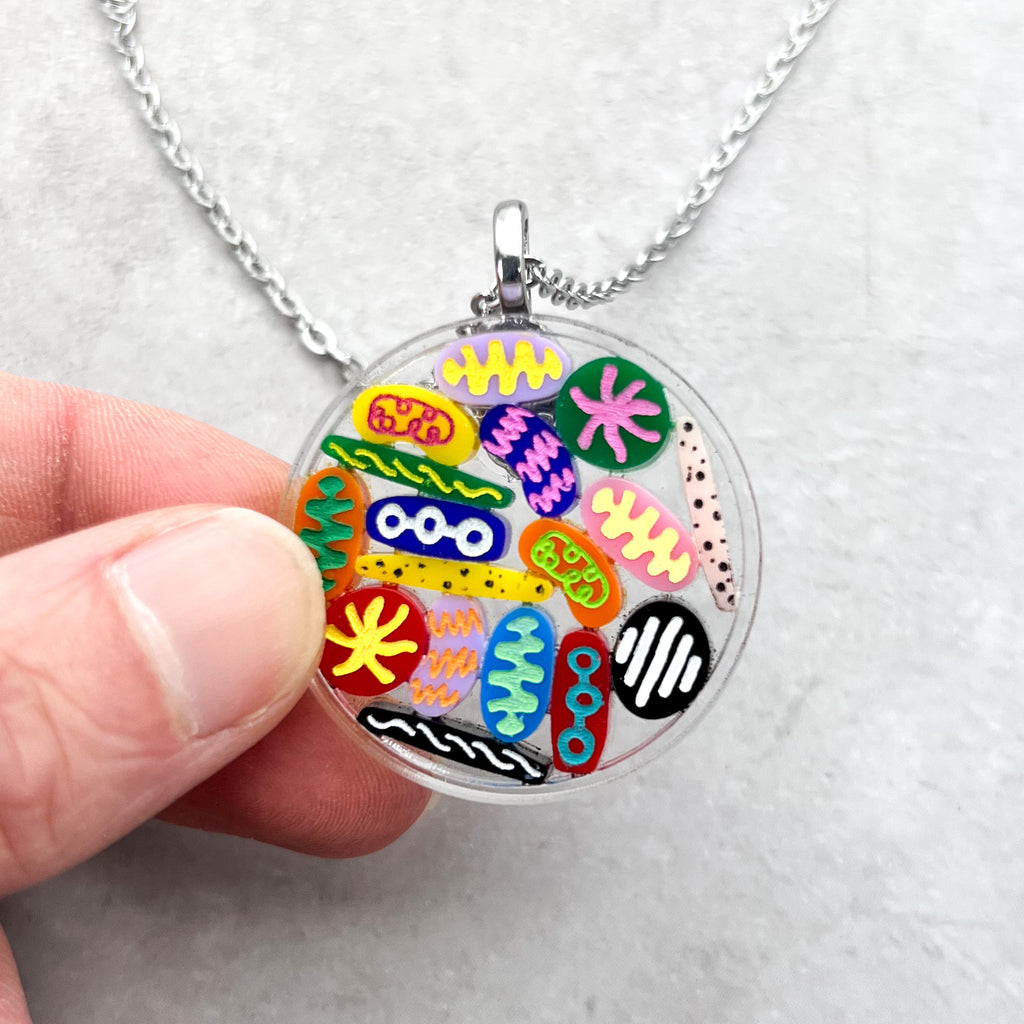 Closeup view of a Petri Dish pendant. Lots of little multicoloured acrylic 'microbes' sit inside an acrylic disc. The pendant hangs from a fine stainless steel acrylic chain.