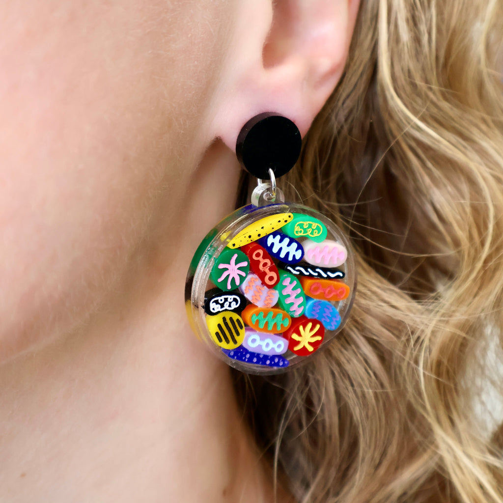 Petri Dish earrings being modelled. Lots of little multicoloured acrylic 'microbes' sit inside an acrylic disc. Earrings hang from black toppers. 