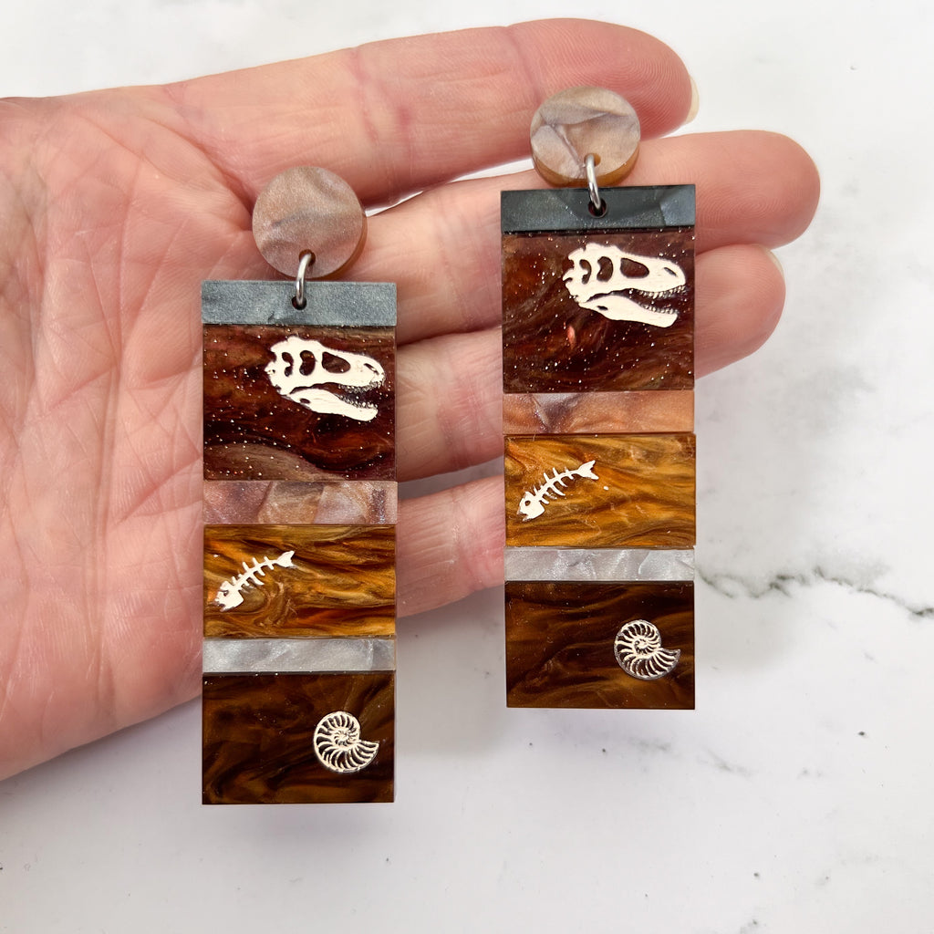 Dangle earrings representing sedimentary layers, comprising 6 strips of different grey and brown acrylics, with a t-rex skull, fish bones and nautilus engraved and handpainted in the larger layers. They hang from brown toppers and are being held up. 