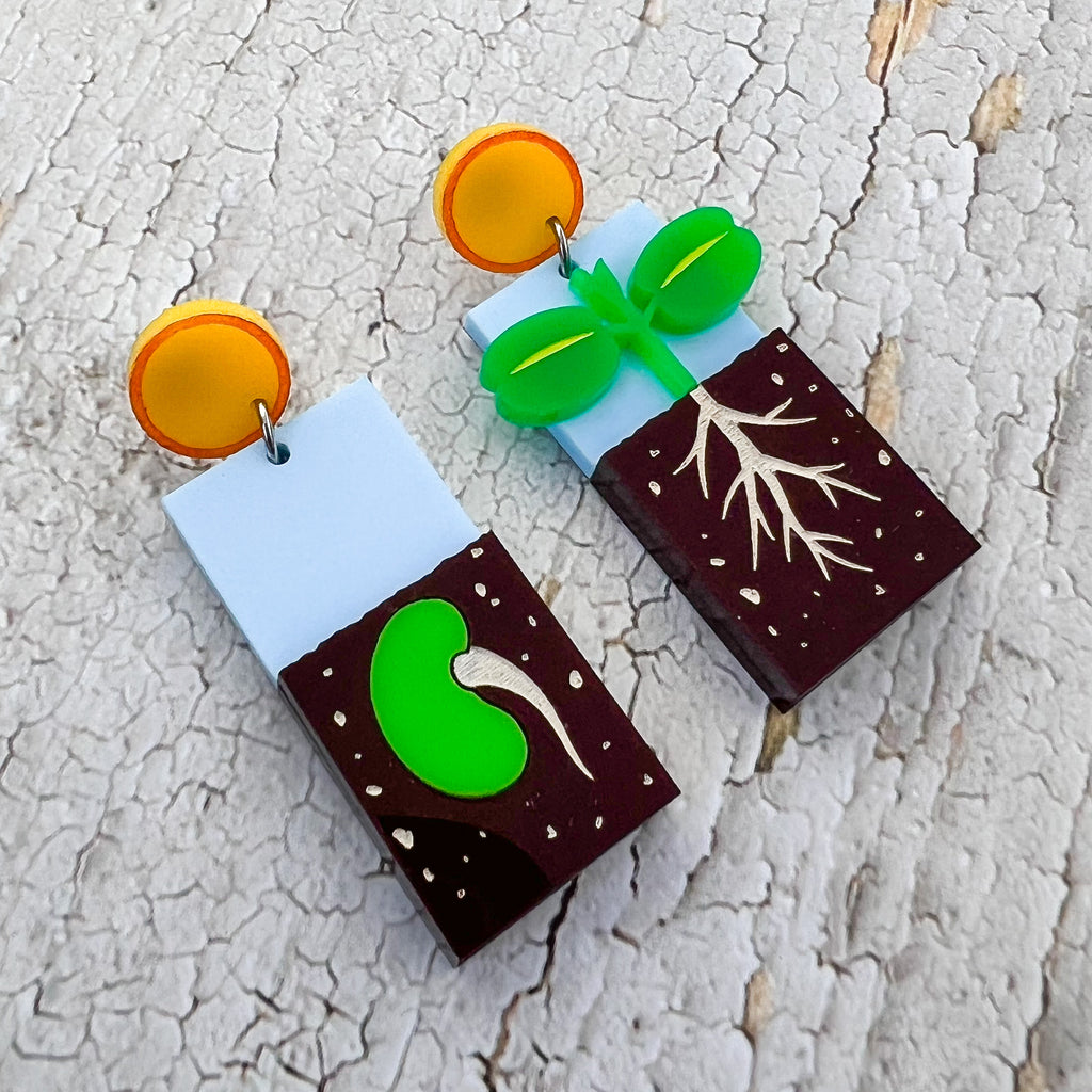 A pair of asymmetrical earrings showing two stages in the germination process. One side shows a seed in the ground while the other side shows a seedling with roots. They hang form yellow 'sun' earring toppers. Laser cut and handpainted from acrylic. 