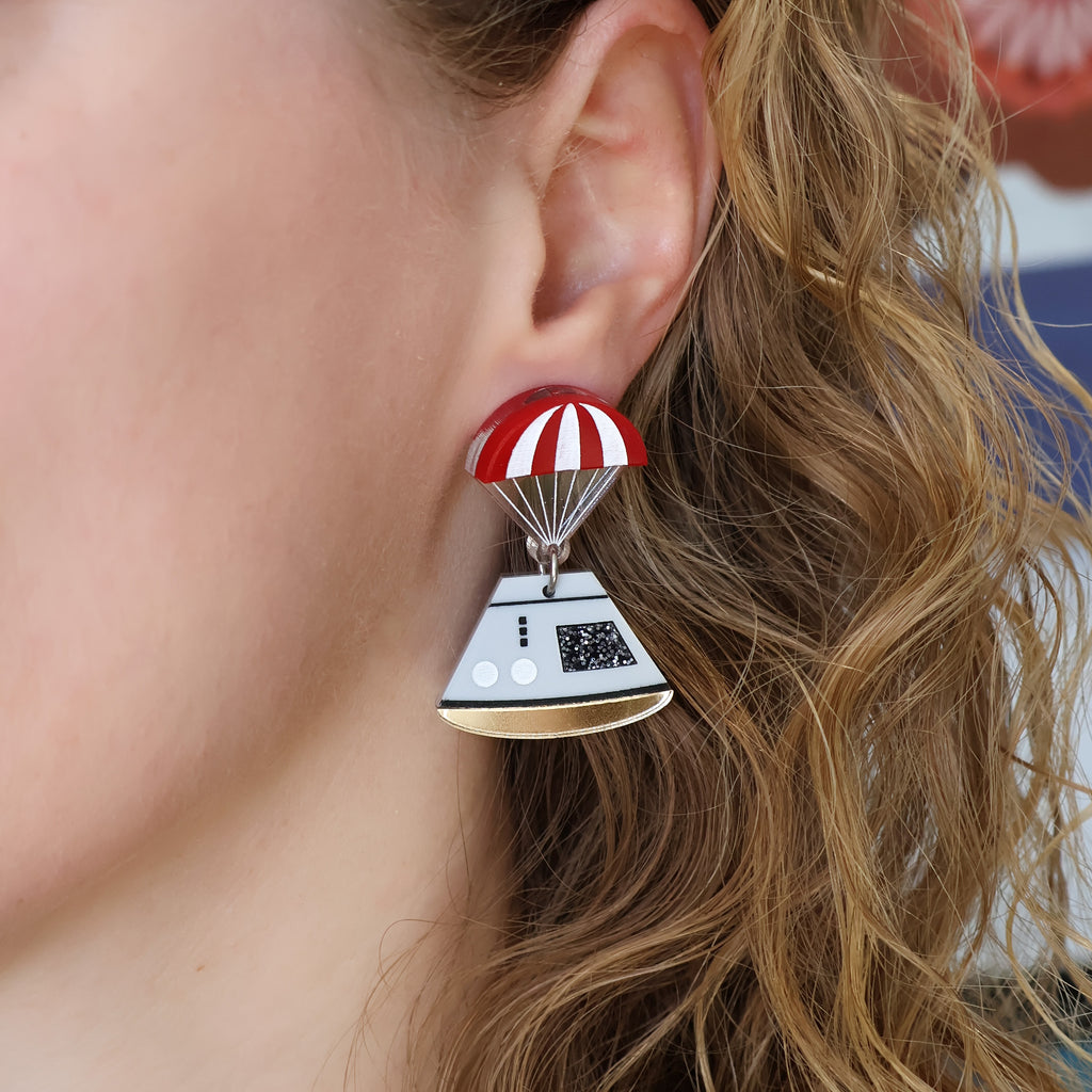 Space capsule earrings, with and red parachute topper and a grey capsule dangling underneath. Laser cut from acrylic. Earrings are being modelled. 