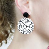 Round white dangle earrings with a black phylogenetic tree designed etched within. Hanging from black round acrylic toppers. Being modelled.