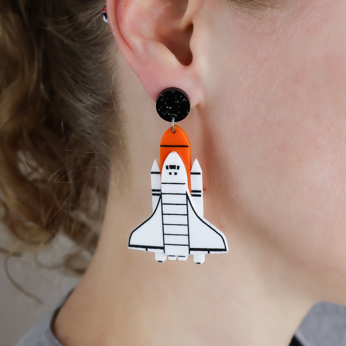 Laser cut acrylic space shuttle earrings in white, orange and black. Being modelled.