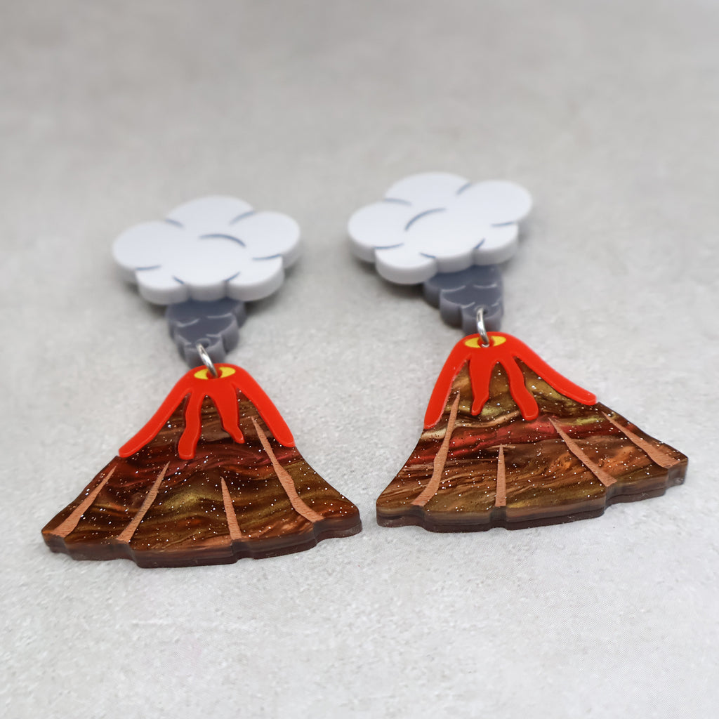 An angled image of acrylic volcano earrings. The tops of the earrings are grey toned ash clouds, and volcanoes with lava flowing out dangle below.