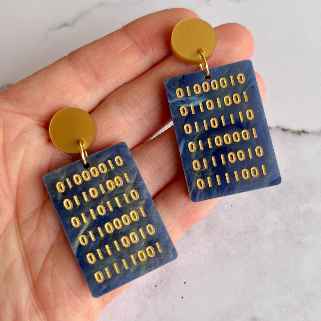 Rectangular earrings in swirly blue acrylic, with an array of gold binary code engraved within. They hang from gold toned acrylic toppers.