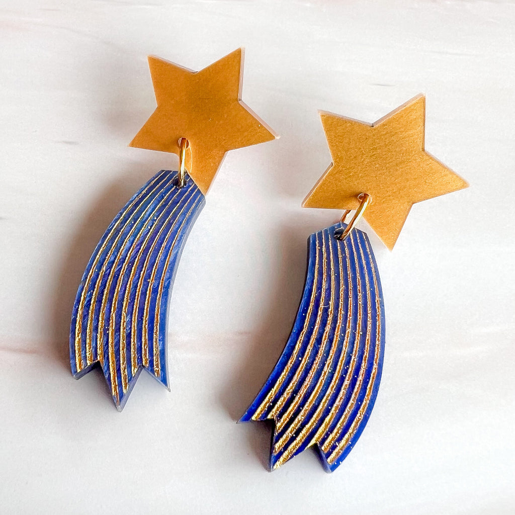 Shooting star earrings, comprised of gold toned acrylic star toppers with blue and gold star trails dangling underneath. 