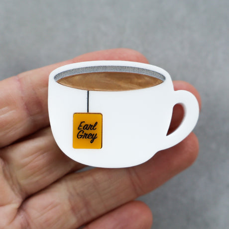 Earl Grey Tea Acrylic Brooch Pin, depicting a teabag hanging from a white cup.  Brooch is being held in a hand.
