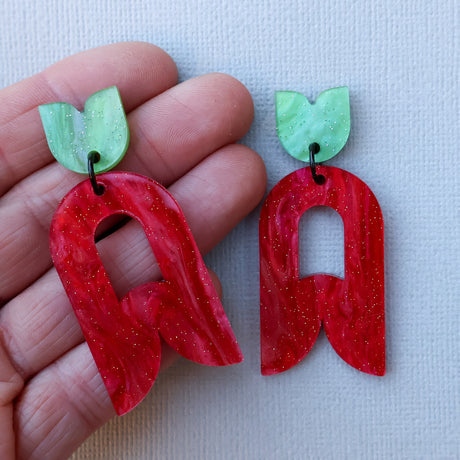 Red and green abstract flower earrings held up in a hand on a light background