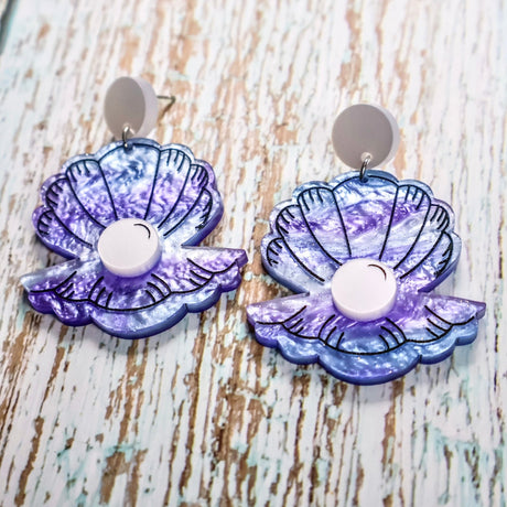 Pair of purple and blue acrylic clam and pearl earrings on a wooden background. Acrylic is swirly with glitter accents. The 'Pearl' is laser cut in a pearlescent acrylic. 