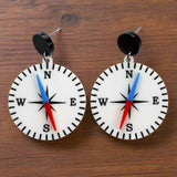 A pair of compass-themed acrylic earrings on a wooden background. The compass is white, with a red and blue needle, and round black toppers. 