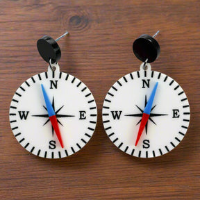 A pair of compass-themed acrylic earrings on a wooden background. The compass is white, with a red and blue needle, and round black toppers. 