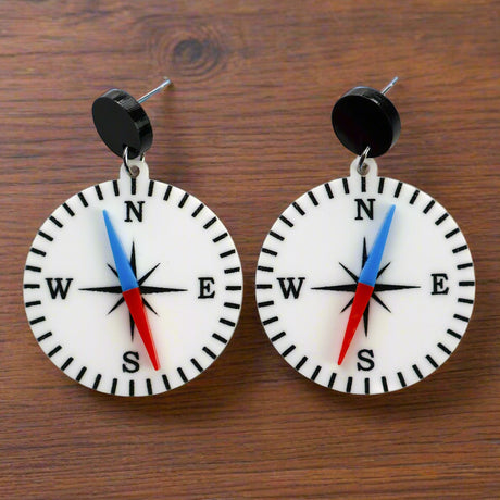 A pair of compass-themed acrylic earrings on a wooden background. The compass is white, with a red and blue needle, and round black toppers. 