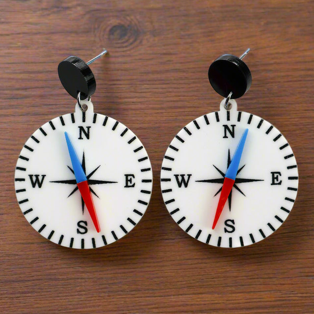 A pair of compass-themed acrylic earrings on a wooden background. The compass is white, with a red and blue needle, and round black toppers. 