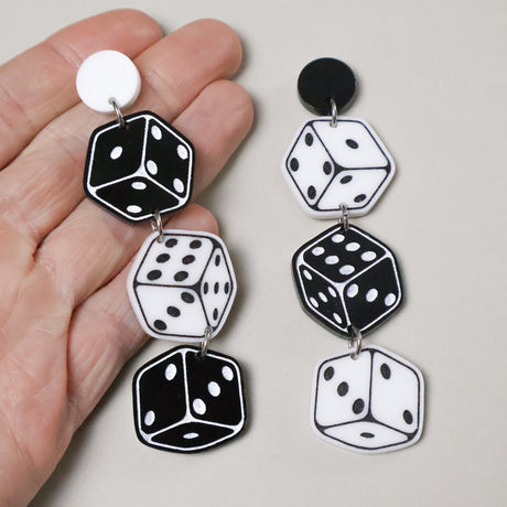 Closeup of a pair of asymmetrical dice themed acrylic earrings. 3 dice hang from round earring toppers in black and white alternating colours. 