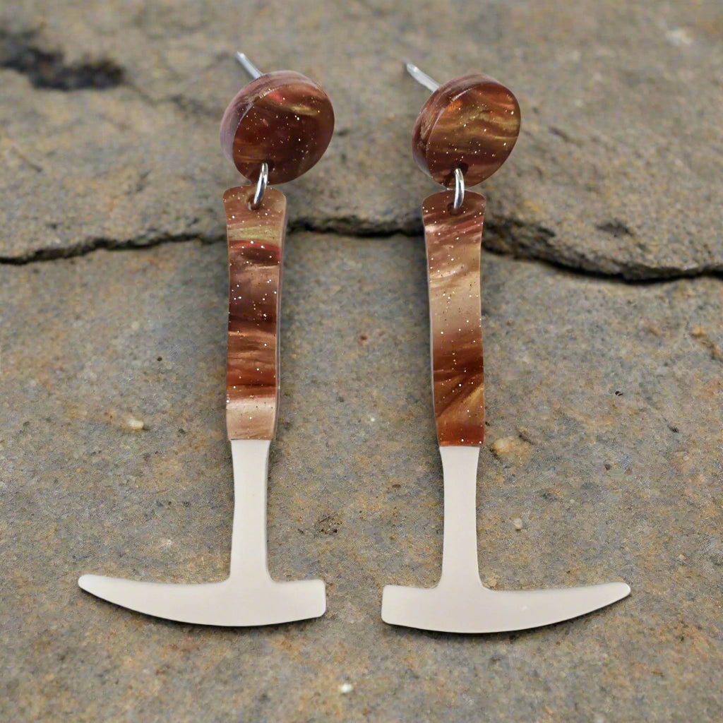 A pair of earrings designed to resemble fossil hammers with a silver pearlescent and brown swirly acrylic finish, attached to surgical steel posts with comfort clutch earring nuts. Laser cut acrylic.