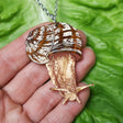 An acrylic snail pendant on a hand with green leafy background. The pendant has a stainless steel chain attached at the top. 